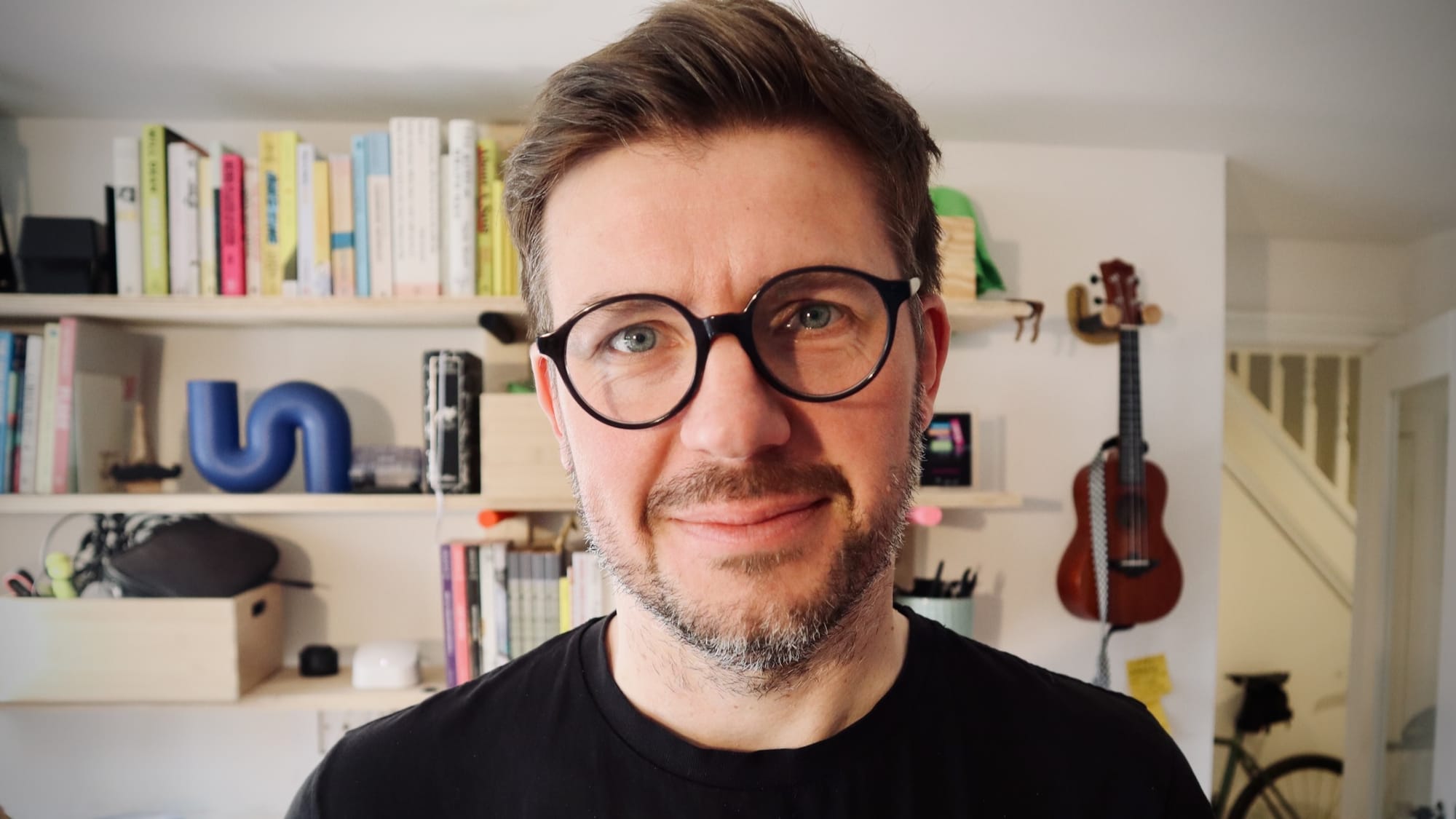 Iain Broome wearing glasses and a beard in front of bookshelves filled with various items like a ukulele and books